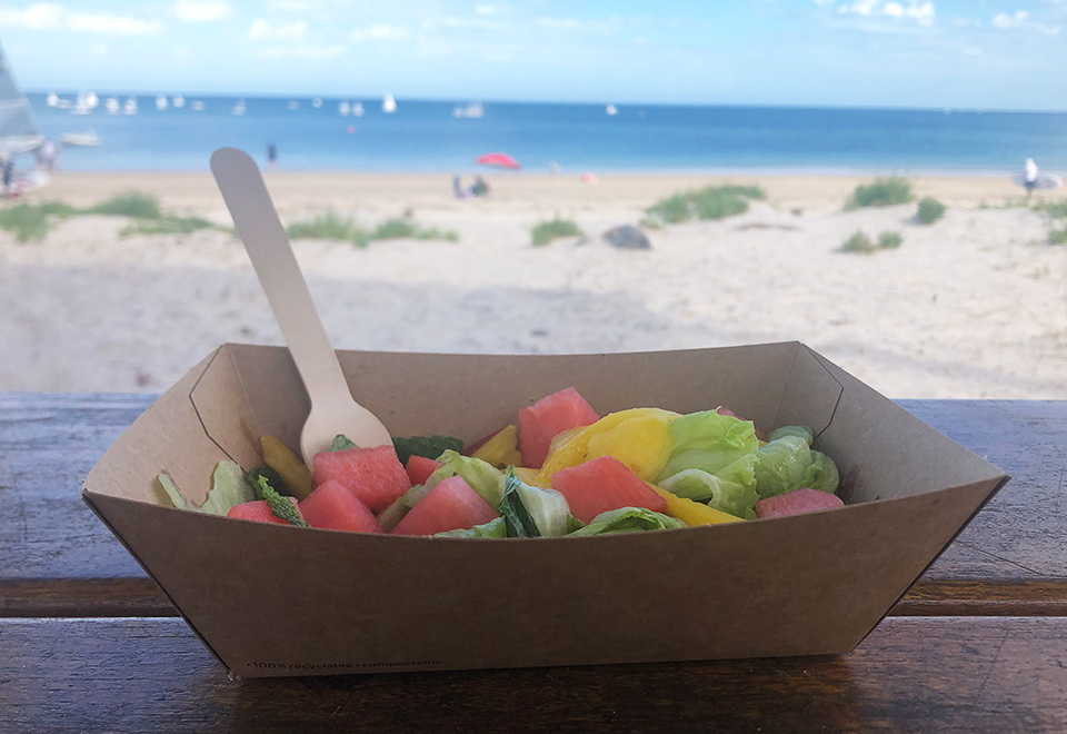 Endura tray filled with fruit salad and pictured with a wooden fork by the beach. 